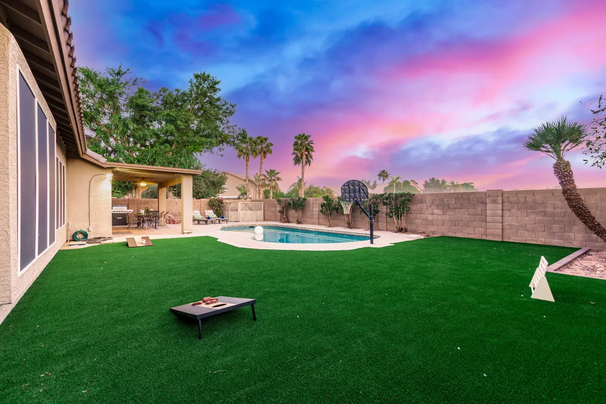 This image captures a lively backyard scene featuring a sparkling swimming pool, a visible basketball hoop attached to the pool, a lush green lawn, and a welcoming patio area. The setup conveys fun, relaxation, and the possibility for both active and leisurely activities.