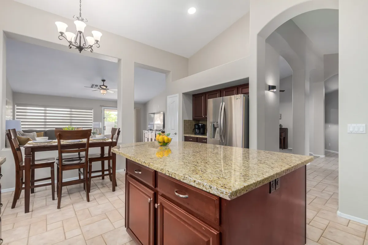 An expansive view of the kitchen showing a large granite island, modern appliances, a wooden dining table, and a stylish chandelier. The space is designed for both cooking and group gatherings, exuding warmth and hospitality.