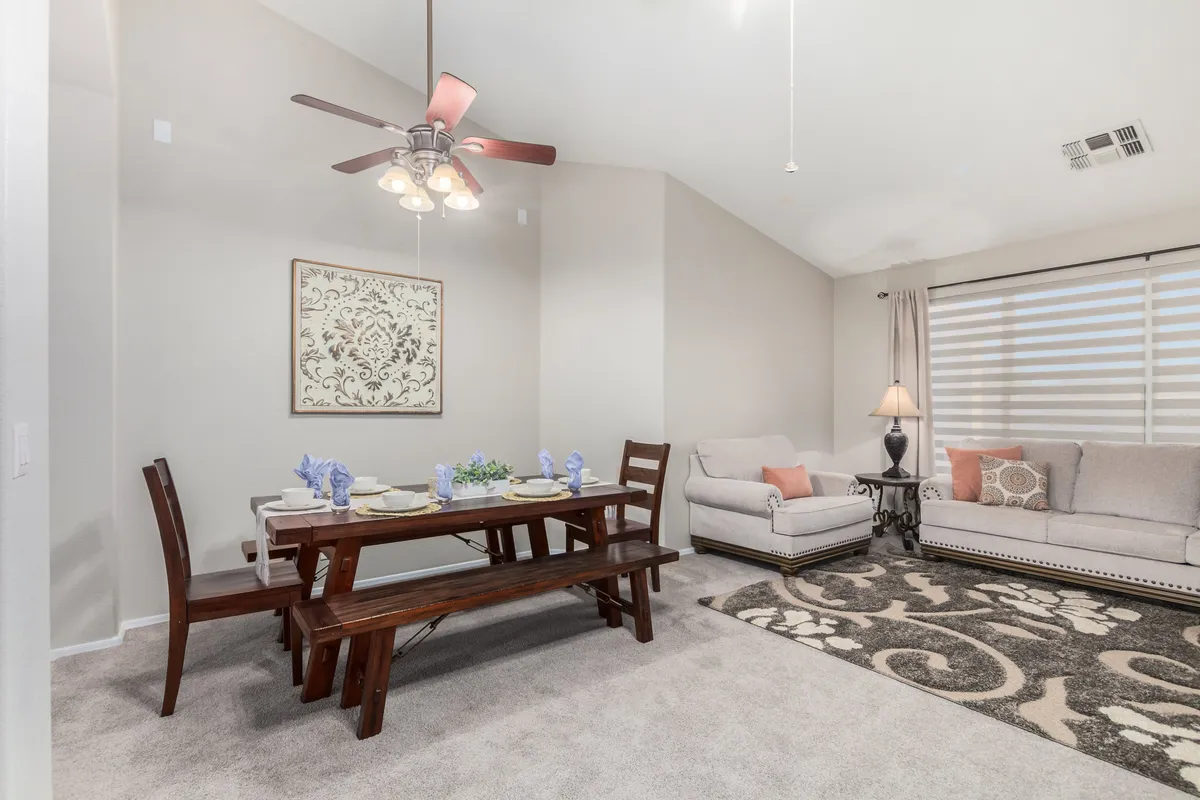A bright and warm interior showing a comfortable sofa, a dining table with place settings, wall art, and an area rug. The space is arranged for social interaction, with the dining area and living area seamlessly connected.