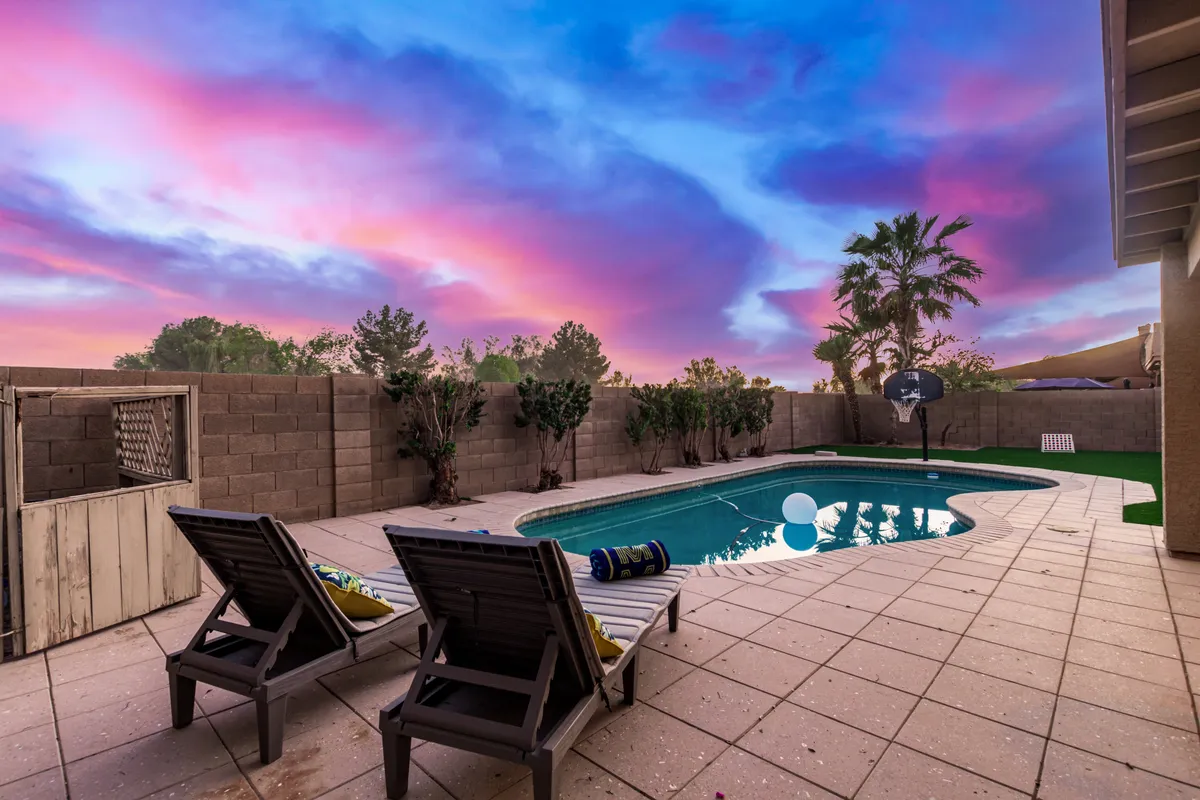 The image captures a tranquil swimming pool surrounded by lounge chairs and palm trees under a colorful, setting sky. The scene suggests peaceful relaxation and scenic beauty, ideal for unwinding after a day of adventure.