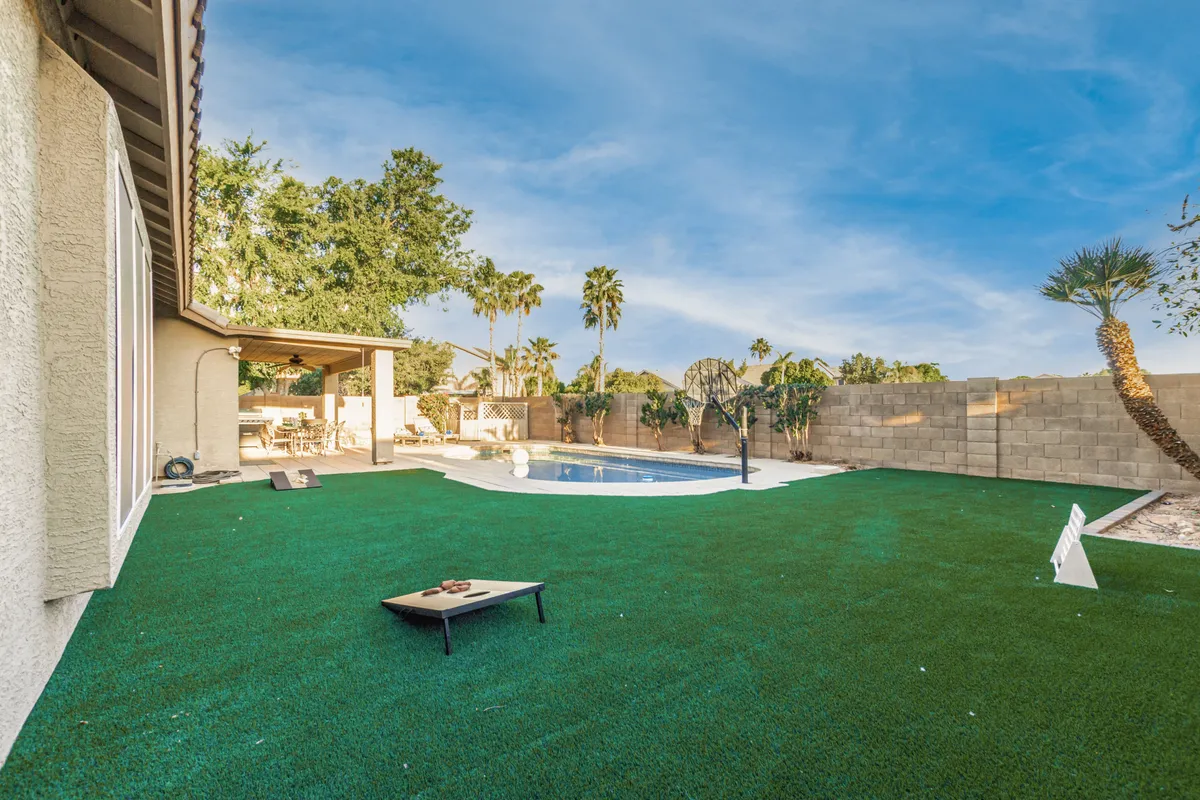 A sun-drenched backyard featuring a sparkling swimming pool surrounded by a spacious lawn area, outdoor seating, and tall palm trees. The scene is inviting and lively, perfect for family games, pool fun, and relaxed gatherings under the Arizona sun.