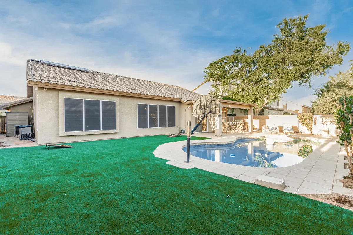A bright exterior shot displaying a swimming pool, a lush green lawn, a patio area equipped for barbecues or dining, and large windows that connect the indoor and outdoor spaces. The scene evokes cheerful gatherings and relaxed moments in the sun.