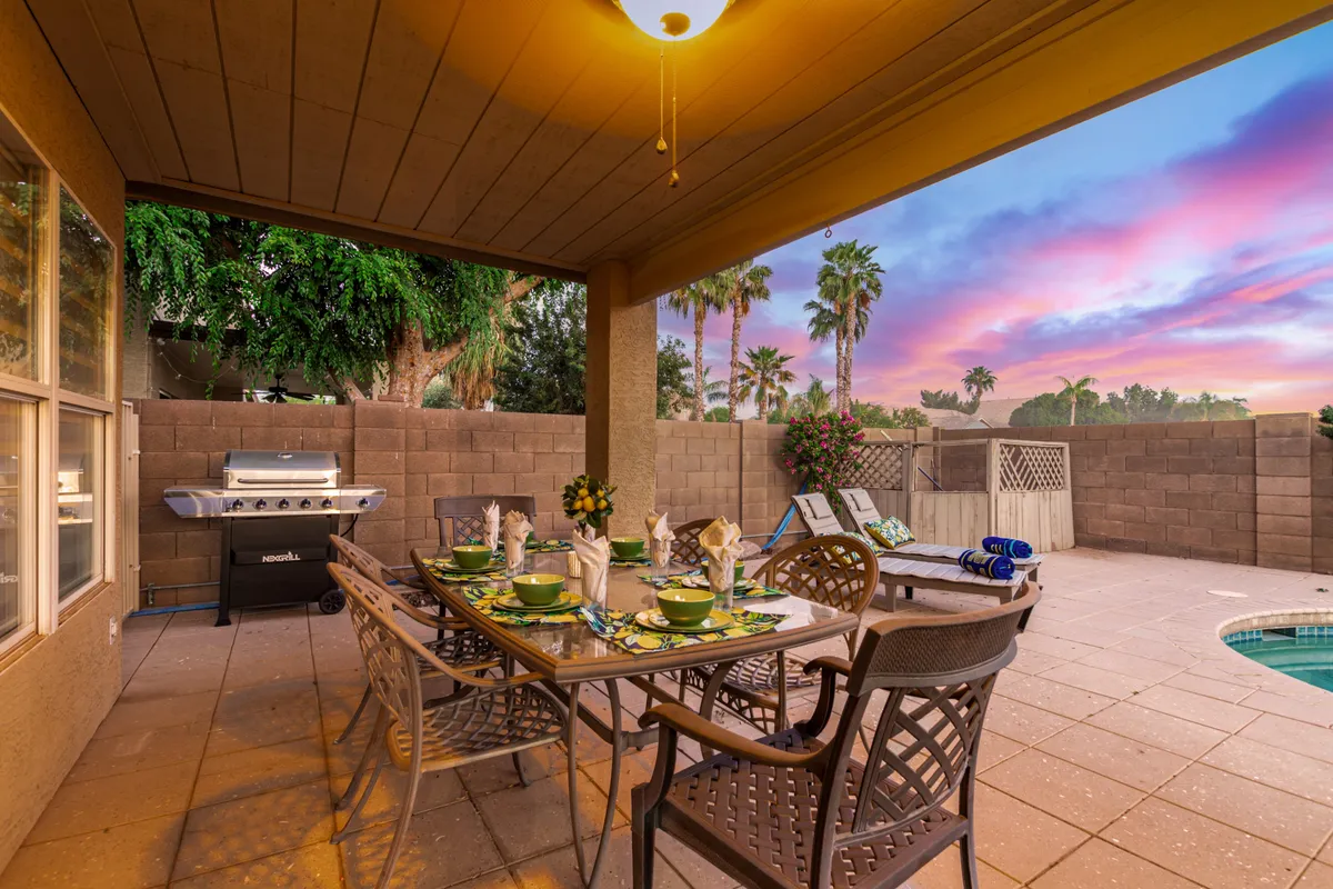 A sunset-lit outdoor dining area is set for a meal, with a grill nearby and the inviting pool glistening in the background. Lush greenery surrounds the space, enhancing its ambiance for alfresco dining and social gatherings.