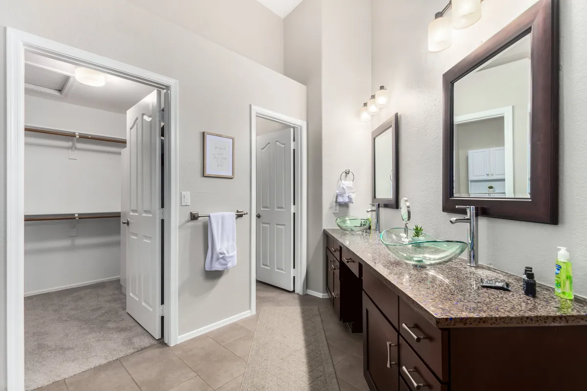 A spacious bathroom scene with a vanity featuring dual glass bowl sinks, large mirrors, and a glimpse into a generous closet space. The area is bright and tranquil, suggestive of a spa-like experience.