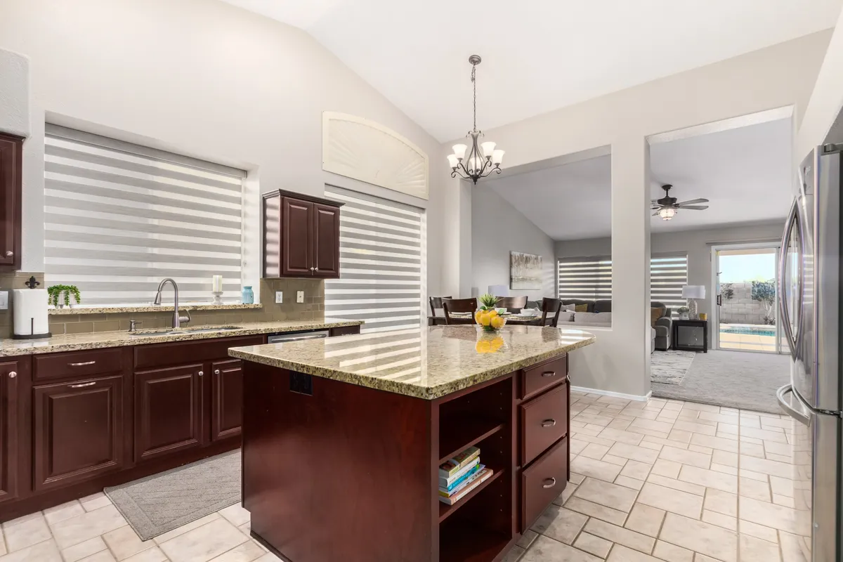 A welcoming kitchen with a prominent granite island at the center, sunlight streaming through the window with blinds, and a dining area visible in the background. The space feels open and is clearly suited for both meal preparation and socializing.