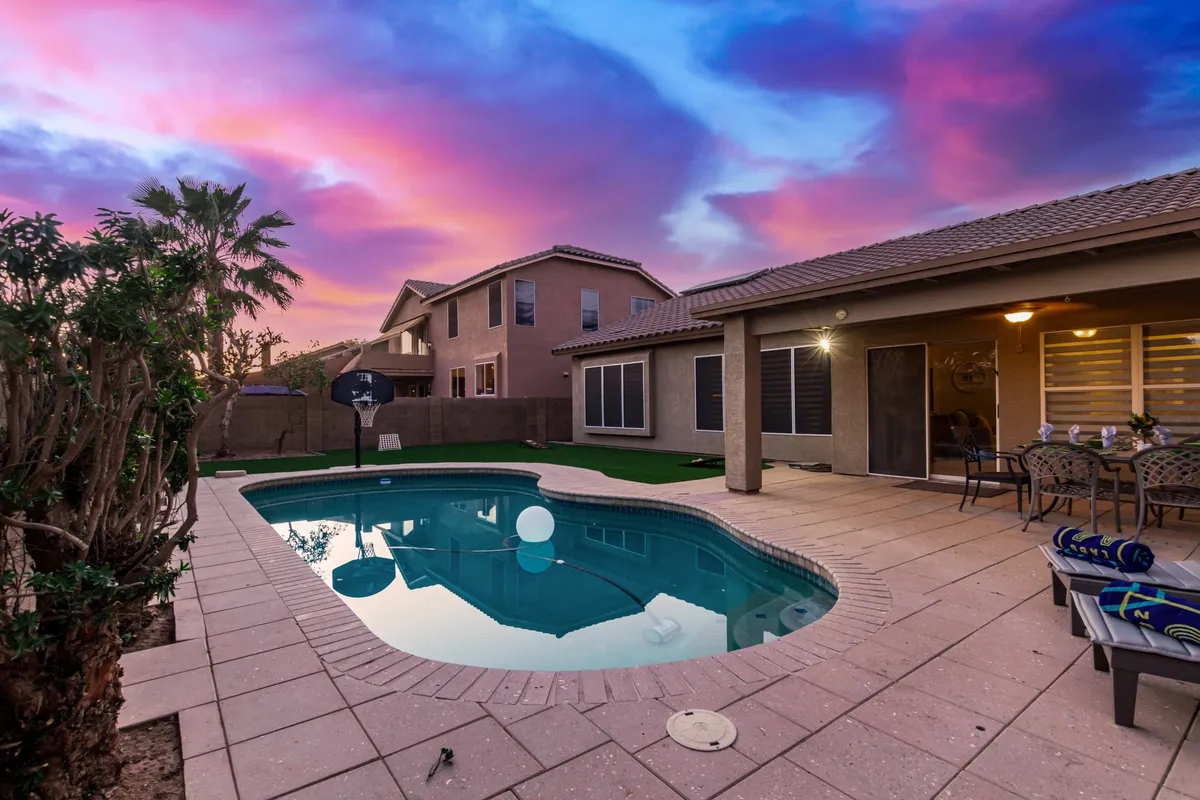 A picturesque backyard scene at sunset shows the shimmering pool, comfortable lounge chairs, and tall palm trees silhouetted against a sky streaked with colorful clouds. The space feels both relaxing and ready for memorable family evenings.